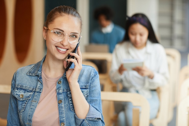 Retrato de sonriente chica atractiva estudiante en anteojos mediante teléfono móvil en la sala de conferencias de la universidad