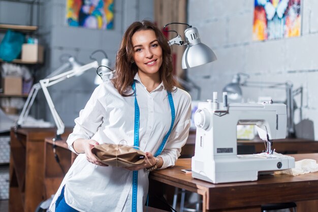 Retrato del sonriente diseñador de moda europeo de pie junto a la máquina de coser sosteniendo un regalo embalado en papel artesanal