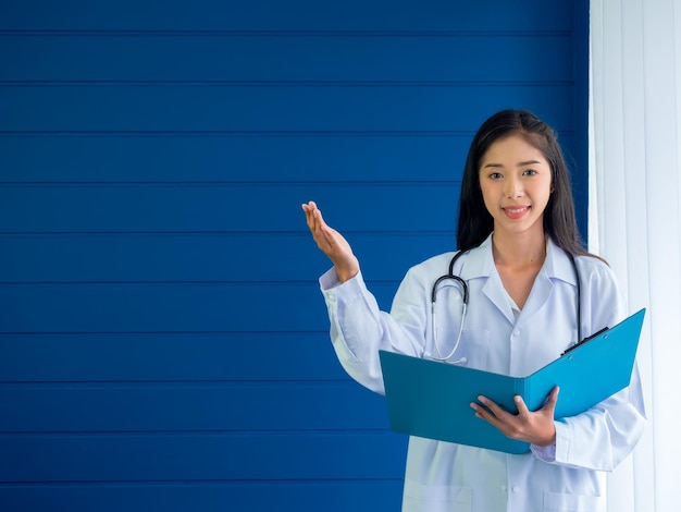 Retrato sonriente de una doctora bastante asiática de pie sobre fondo azul sosteniendo una carpeta de documentos y mostrando un gesto de mano en un espacio en blanco Practicante joven segura de sí misma sosteniendo una carpeta de documentos