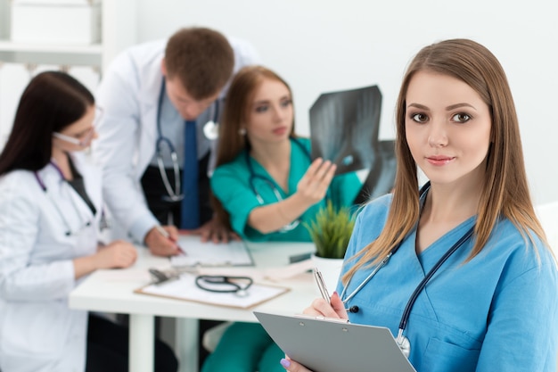 Retrato de sonriente doctora en medicina sosteniendo una carpeta con documentos con sus tres colegas trabajando. Concepto de salud y medicina.