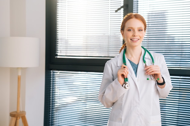 Retrato de sonriente doctora de pie sobre el fondo de la ventana en un día soleado en el consultorio médico