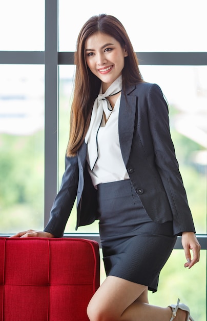 Foto retrato de sonriente empresaria asiática o secretaria vistiendo ropa elegante apoyado en el sofá en el pasillo de la oficina y mirando a la cámara