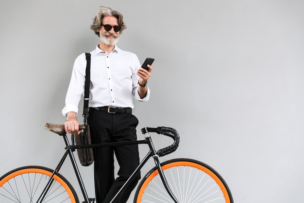 Retrato de sonriente empresario barbudo en gafas de sol con teléfono móvil mientras está de pie con bicicleta sobre pared gris al aire libre