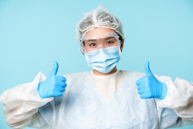 Retrato de sonriente enfermera o médico asiático, vistiendo equipo de protección personal, mostrando los pulgares hacia arriba, recomendando, de pie sobre fondo azul.