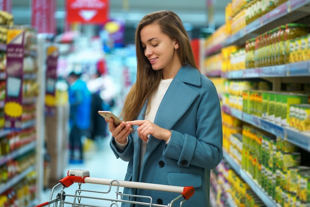 Retrato de una sonriente feliz atractiva joven comprador con carro en el pasillo de la tienda con la lista de compras en el teléfono inteligente durante la compra de alimentos