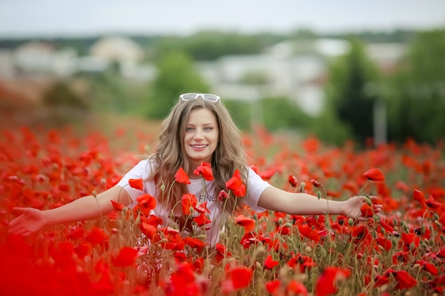 El retrato sonriente feliz hermoso de la muchacha adolescente con las flores rojas en la cabeza que goza en amapolas coloca el fondo de la naturaleza. Maquillaje y peinado rizado. Estilo de vida.