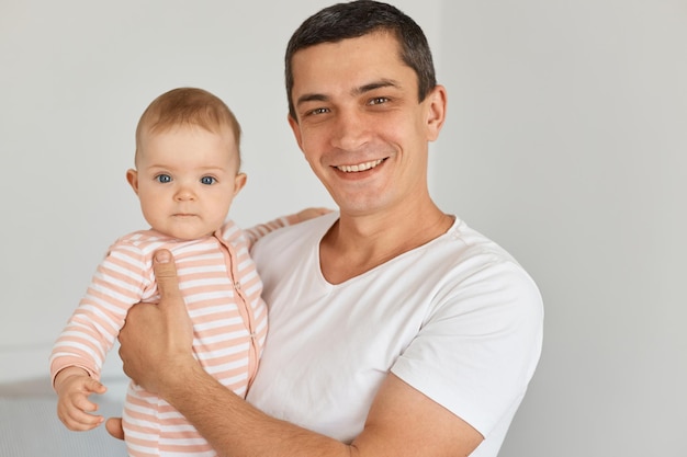 Retrato de sonriente feliz morena hombre vestido con camiseta blanca casual sosteniendo a su linda hija pequeña, posando en casa en la habitación de luz, mirando a cámara, expresando emociones positivas.