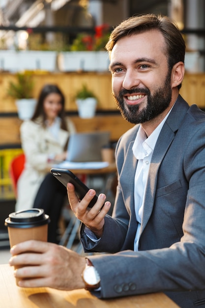 Retrato de sonriente hombre sin afeitar vestido con chaqueta bebiendo café para llevar y utilizando un teléfono móvil mientras está sentado en la cafetería