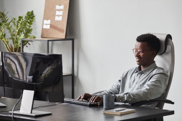 Retrato de sonriente hombre afroamericano con computadora mientras está sentado en un escritorio en la oficina, concepto de desarrollador de software, espacio de copia