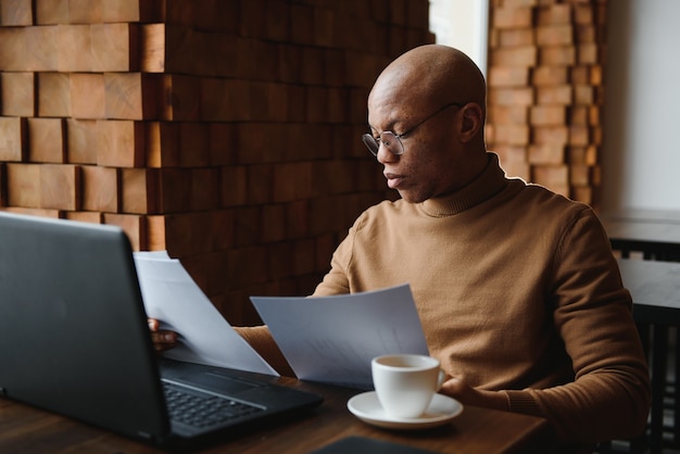 Retrato de sonriente hombre afroamericano en gafas sentarse en un escritorio en la oficina trabajando en equipo portátil