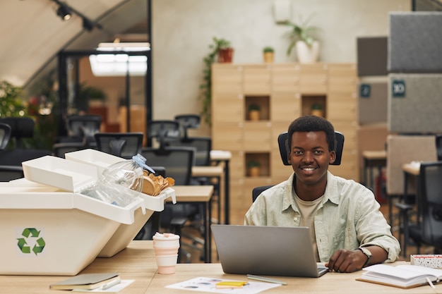 Retrato de sonriente hombre afroamericano con portátil mientras está sentado por contenedores de clasificación de residuos en la oficina moderna, espacio de copia