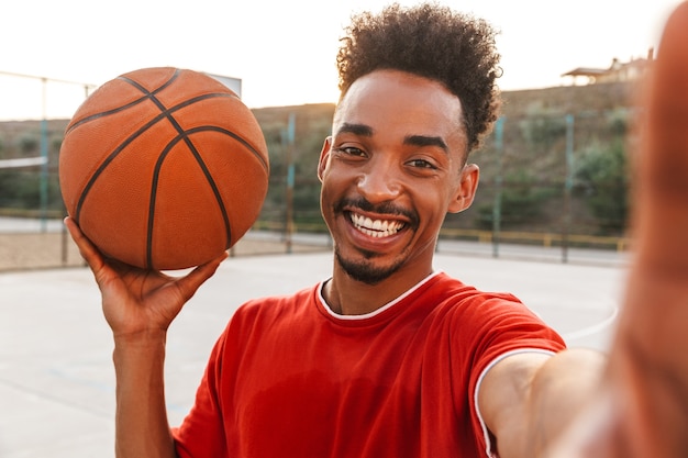 Retrato de sonriente hombre afroamericano sosteniendo la pelota y tomando selfie, mientras juega baloncesto en el patio de recreo al aire libre