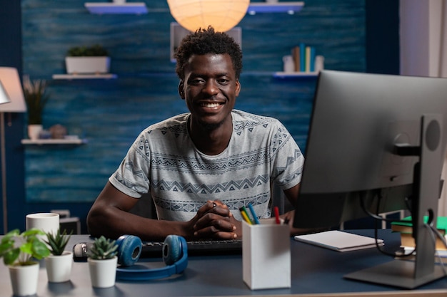 Retrato de sonriente hombre afroamericano usando la computadora mientras trabaja de forma remota desde casa en un proyecto de marketing. Empleado joven analizando gráfico financiero sentado en el escritorio con tecnología de oficina de negocios