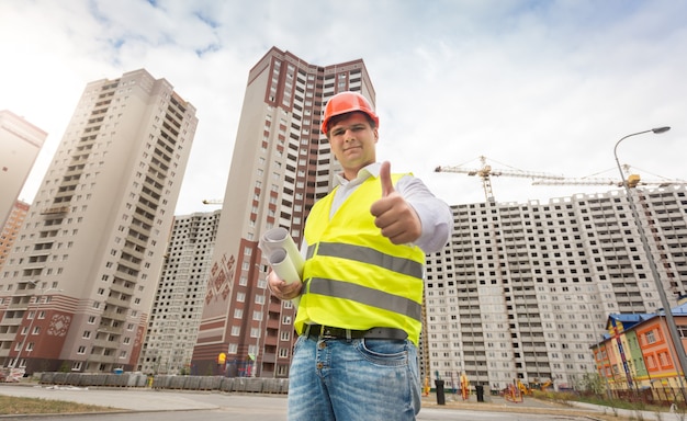 Foto retrato de sonriente ingeniero de construcción de pie en el sitio de construcción y sosteniendo los pulgares hacia arriba