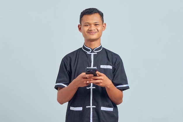 Retrato de sonriente joven asiático vistiendo uniforme de karate mediante teléfono móvil sobre fondo gris