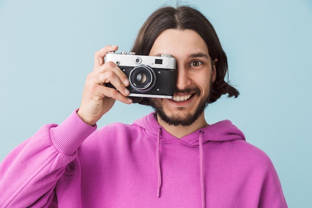 Retrato de un sonriente joven barbudo morena hombre vestido con sudadera con capucha que se encuentran aisladas sobre la pared azul, tomando fotografías con cámara de fotos