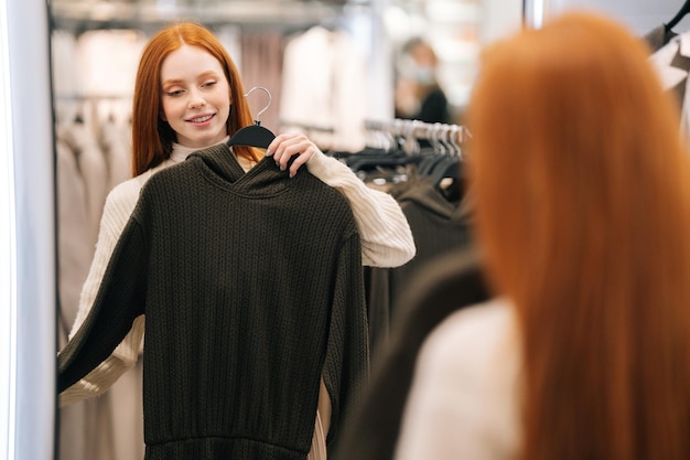 Retrato de sonriente joven comprador eligiendo ropa y mirando al espejo en la tienda de ropa