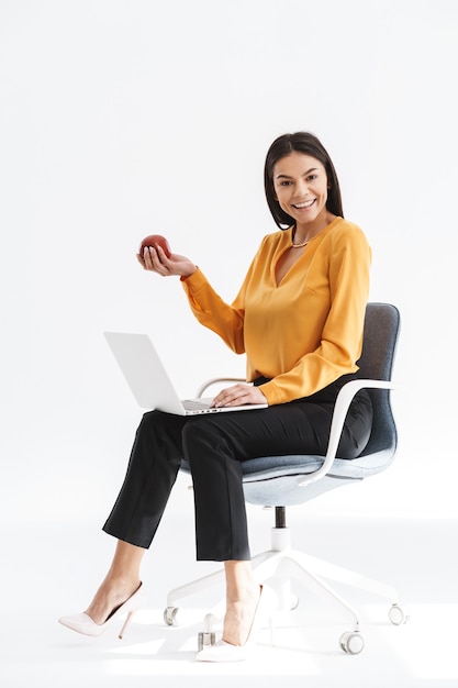 Retrato de sonriente joven empresaria vestida con ropa elegante sentado en una silla y usando la computadora portátil mientras trabaja en la oficina brillante