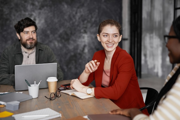 Retrato de sonriente joven empresaria vistiendo chaqueta roja durante la reunión con compañeros de trabajo en la oficina, espacio de copia