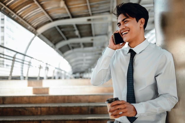 Retrato de un sonriente joven empresario asiático mediante teléfono móvil en la ciudad