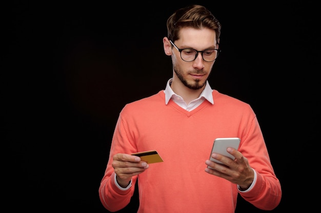 Retrato de sonriente joven guapo en anteojos con aplicación de teléfono inteligente mientras se comprueba el saldo de la tarjeta de crédito