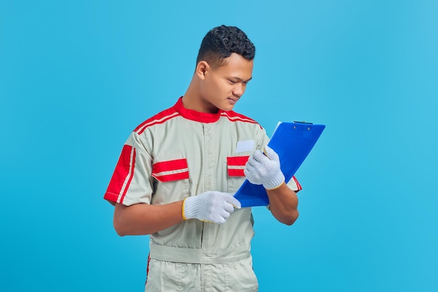 Retrato de sonriente joven mecánico asiático mirando notas en el portapapeles sobre fondo azul.