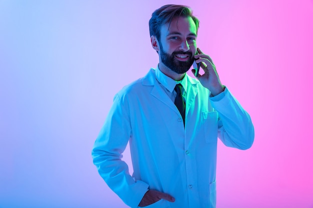 Retrato de un sonriente joven médico con uniforme que se encuentran aisladas sobre neblina rosa-azul, hablando por teléfono móvil