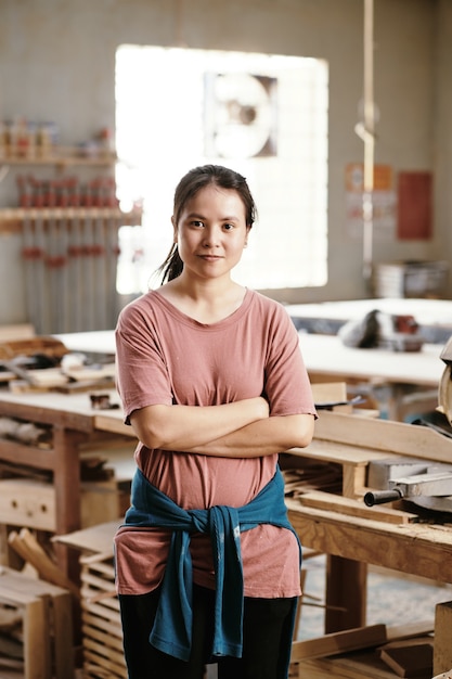 Retrato de sonriente joven mujer asiática segura de pie en el taller de carpinteros en el que está trabajando