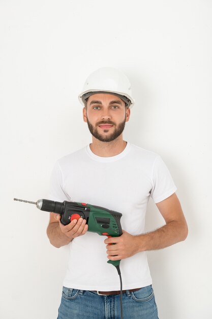 Retrato de sonriente joven reparador barbudo en casco con destornillador inalámbrico y taladro contra la pared blanca