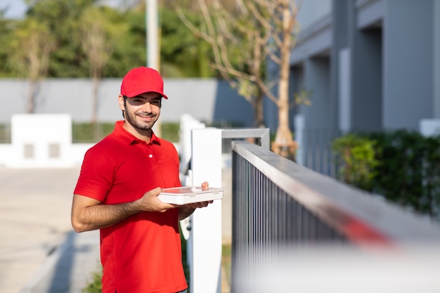 Retrato Sonriente joven repartidor en uniforme rojo sosteniendo una caja en frente de la casa.