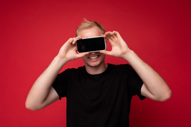Retrato de sonriente joven rubio guapo con camiseta negra de pie aislado en superficie roja sosteniendo teléfono móvil mostrando smartphone en mano con pantalla vacía para maqueta