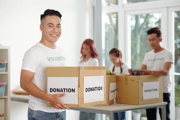 Retrato de sonriente joven sosteniendo caja de donación, sus compañeros de trabajo clasificando ropa y comestibles en segundo plano.