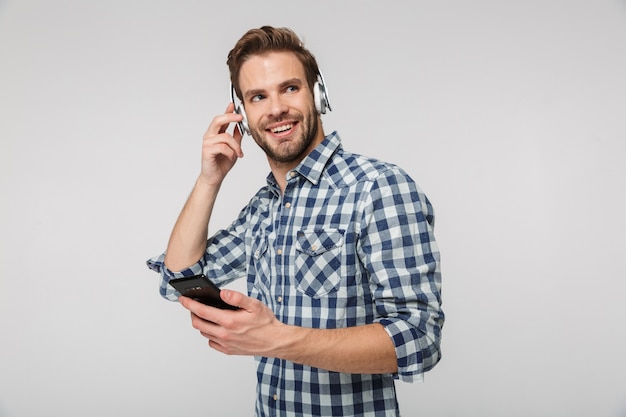 Retrato de sonriente joven usando audífonos con teléfono celular aislado sobre pared blanca