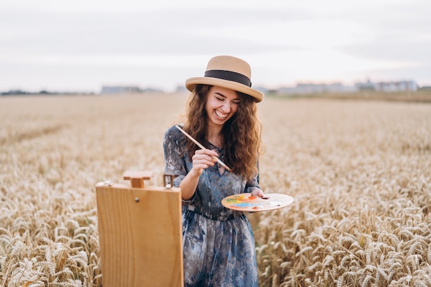 Retrato de sonriente mujer artista con cabello rizado en sombrero. La mujer dibuja un paisaje en un campo de trigo. Copia espacio