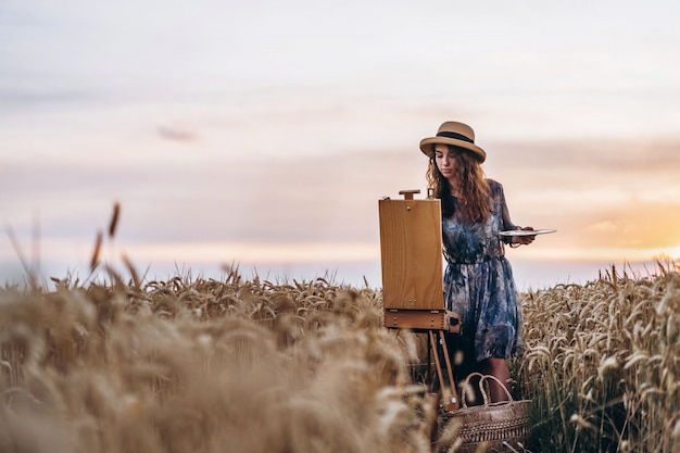 Retrato de sonriente mujer artista con cabello rizado en sombrero. La mujer dibuja un paisaje en un campo de trigo. Copia espacio