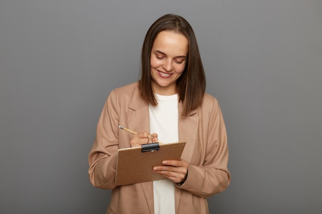 Retrato de una sonriente mujer caucásica de cabello oscuro con chaqueta beige de pie con lápiz y portapapeles escribiendo para hacer la lista de su trabajo posando aislada en un fondo gris