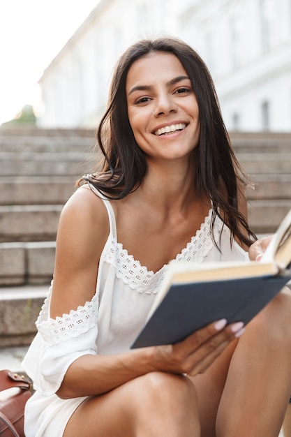 Retrato, de, sonriente, mujer joven, vestido, en, verano, desgaste, libro de lectura, mientras, sentado, en, escaleras, aire libre, wih, taza de papel