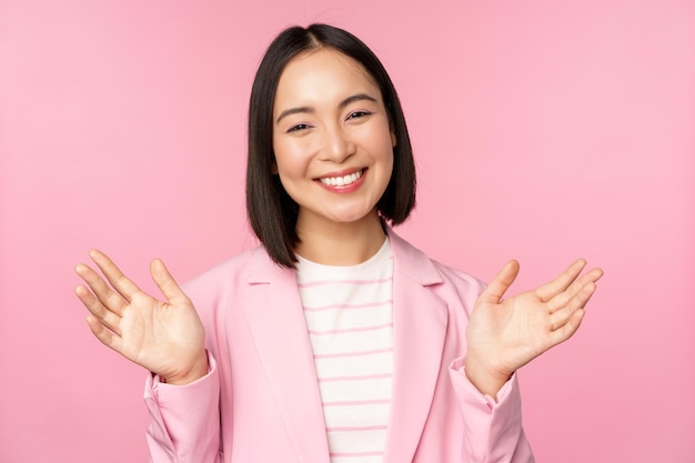 Retrato de una sonriente mujer de negocios de oficina asiática levantando las manos saludando y luciendo feliz posando en traje contra un fondo rosa