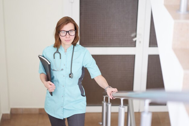 Retrato sonriente de mujer pelirroja blanca médico con estetoscopio en el cuello levantarse de las escaleras