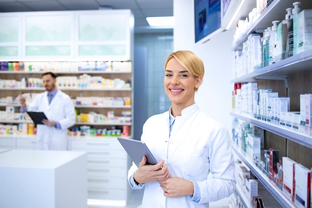 Retrato de sonriente mujer rubia farmacéutica de pie en la farmacia o droguería junto al estante con medicamentos y sosteniendo la tableta.