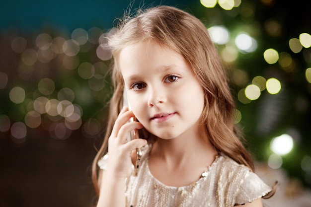 Retrato de una sonriente niña de pelo largo en vestido encendido de luces de Navidad. Niña hablando por teléfono. Año Nuevo y Navidad.