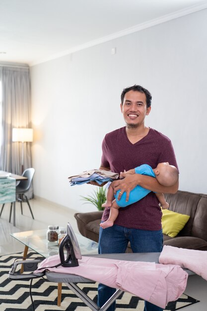 Retrato de sonriente padre asiático planchando su ropa mientras sostiene a su bebé en su mano