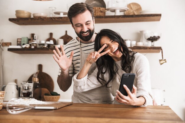 Retrato de sonriente pareja hombre y mujer de 30 años vistiendo delantales abrazándose juntos y sosteniendo el teléfono inteligente mientras se cocina en la cocina de casa