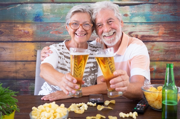 Retrato de sonriente pareja senior brindando con cervezas sentado en la mesa de madera mirando a la cámara