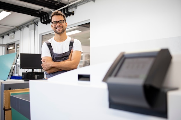 Retrato de sonriente trabajador de impresión de pie junto a la computadora a la máquina de placa en la imprenta.