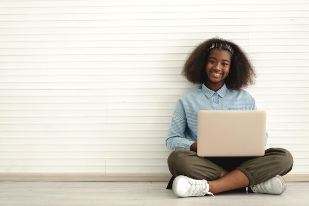 Retrato de sonrisa negro afroamericano lindo estudiante niña sentada en el piso usando tecnología computadora portátil escribiendo en el teclado aprender y estudiar sobre fondo de pared blanca