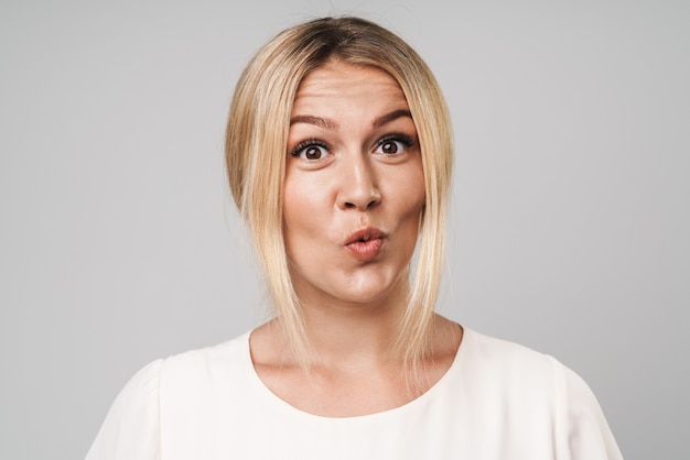 Retrato de una sorprendida sorprendida feliz hermosa joven increíble mujer rubia posando aislada sobre pared gris vestida con camiseta blanca básica