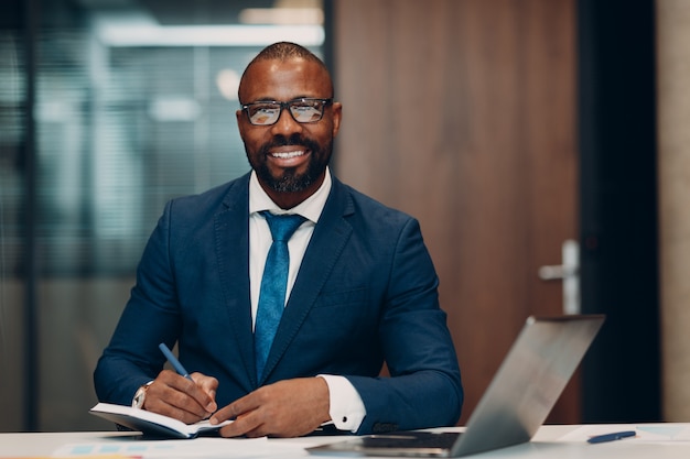 Retrato sorridente empresário afro-americano assina contrato, sente-se à mesa para uma reunião no escritório com caderno com caneta e laptop