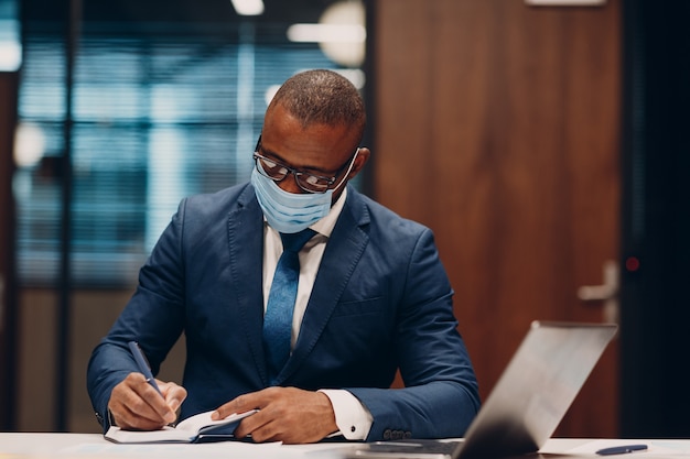 Retrato sorridente empresário afro-americano com máscara protetora médica, sente-se à mesa para uma reunião no escritório com caderno com caneta e laptop
