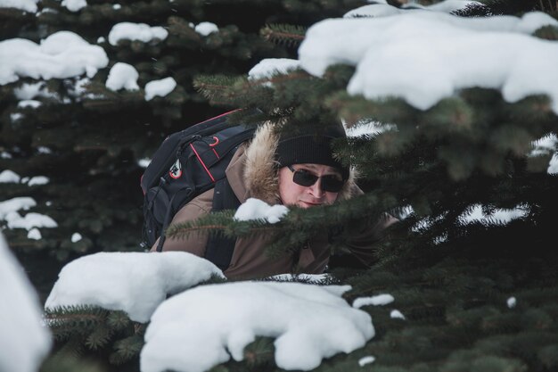 Retrato sorridente jovem brutal ao ar livre entre árvores de Natal cobertas de neve no inverno na floresta. Turista com óculos e mochila se perdeu em uma área desconhecida. Caminhadas loucas de adolescentes